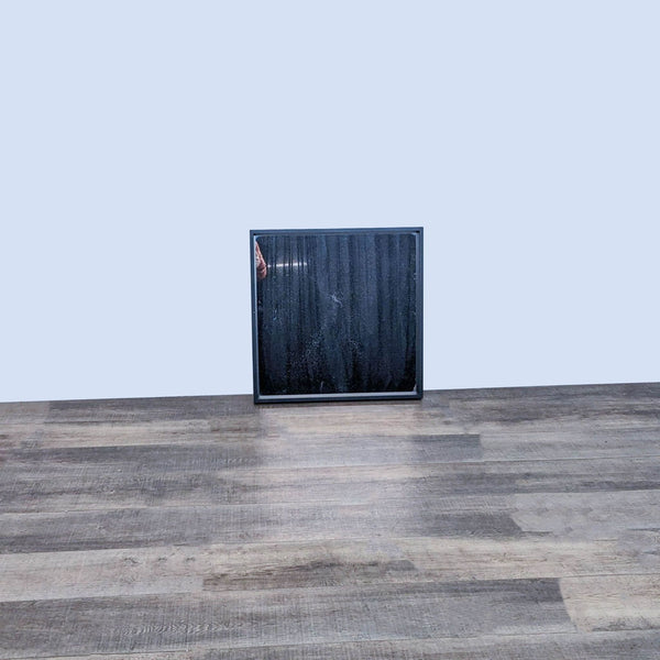 Square wall mirror with a black frame on a wooden floor.