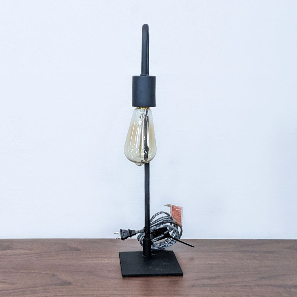 Minimalist black metal lamp with a clear bulb and cord on a wooden table.