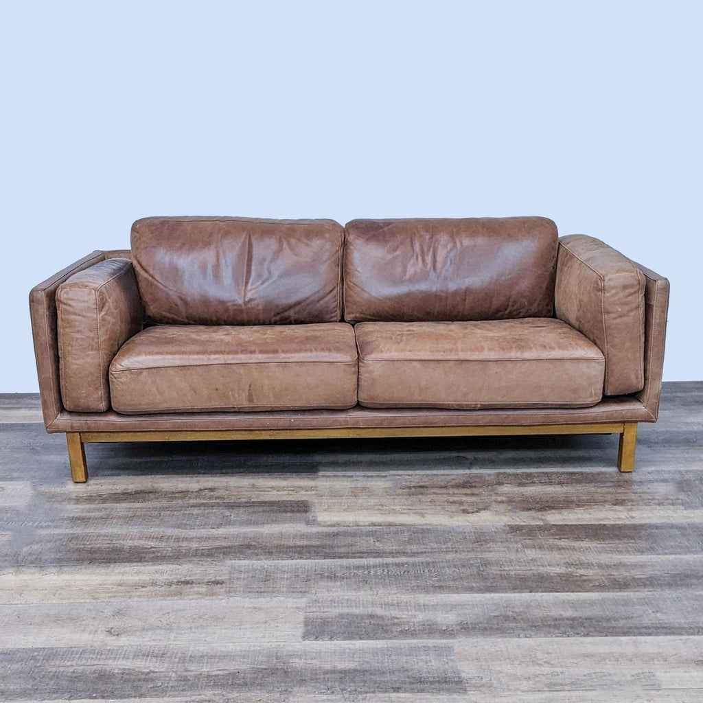 Brown leather sofa with double arms and wooden legs on a hardwood floor.