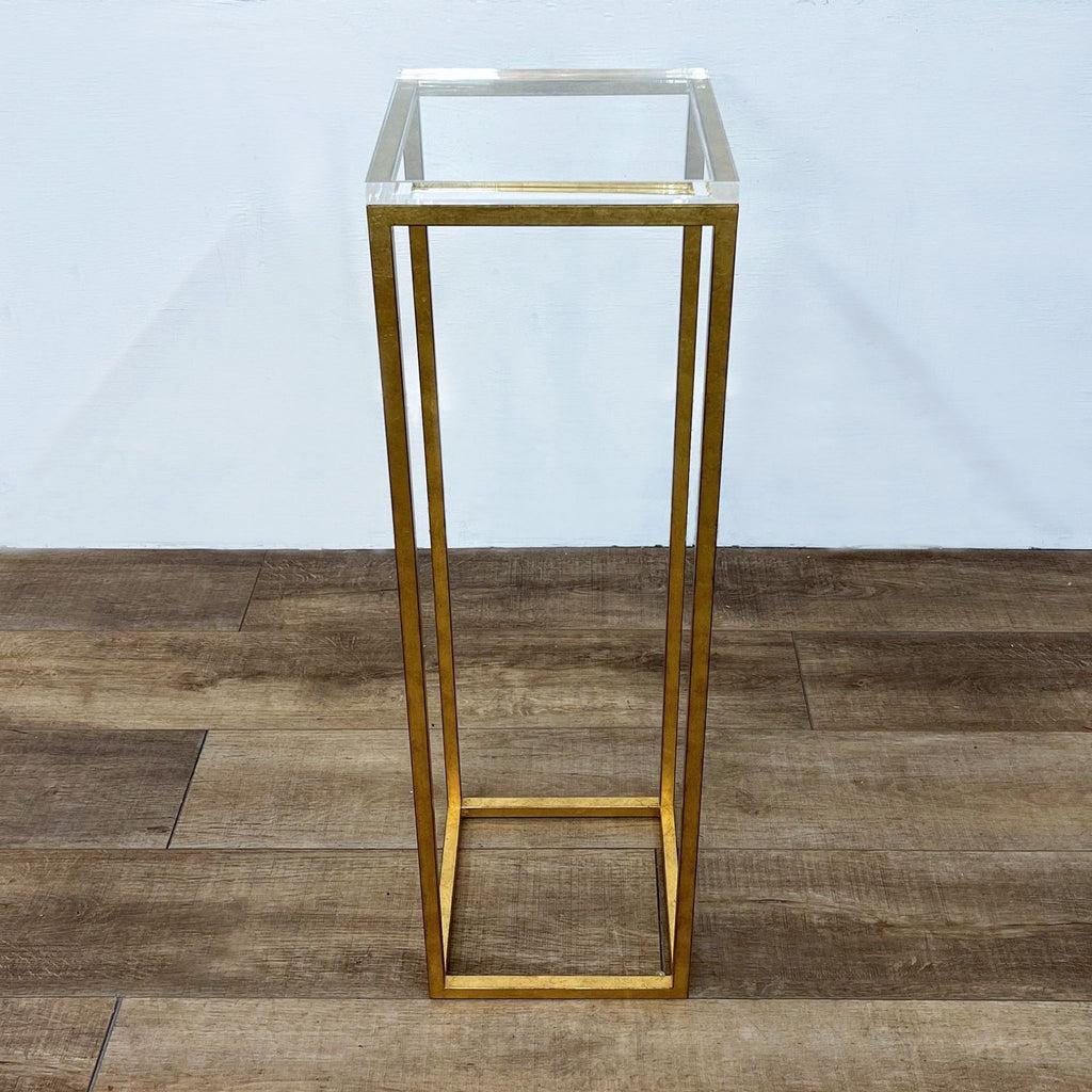 A tall, gold-colored metal stand with a clear glass top on a wooden floor.