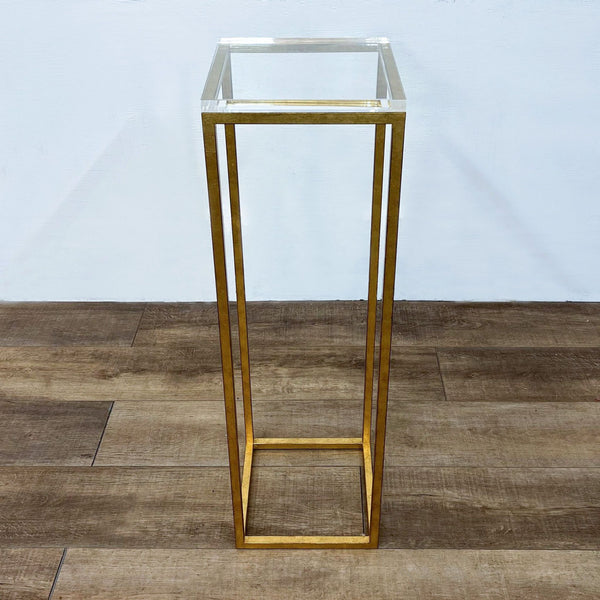 A tall, gold-colored metal stand with a clear glass top on a wooden floor.