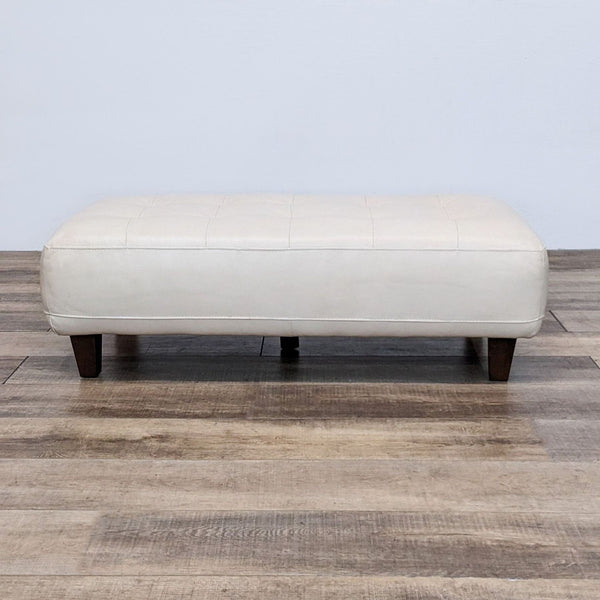 Modern leather ottoman with tufted top and dark wooden legs on a wooden floor.