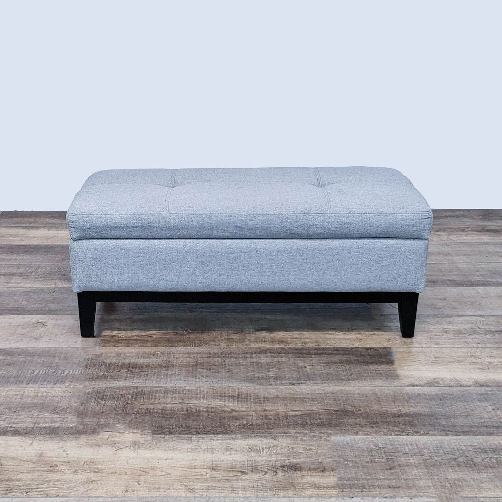 Front view of gray fabric ottoman, tufted top, dark wood legs.