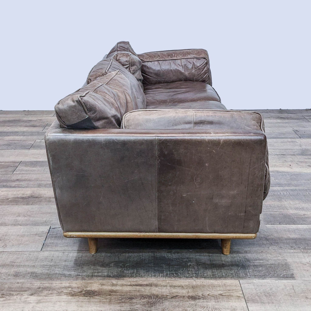 Side view of a brown leather sofa with cushion arms on wooden legs.