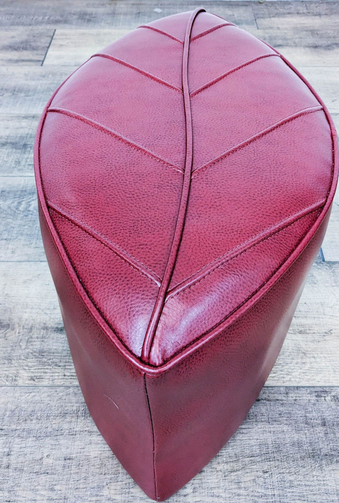 Red leaf-shaped ottoman, leather look, on wooden floor. Category: Stools, Ottomans & Benches.