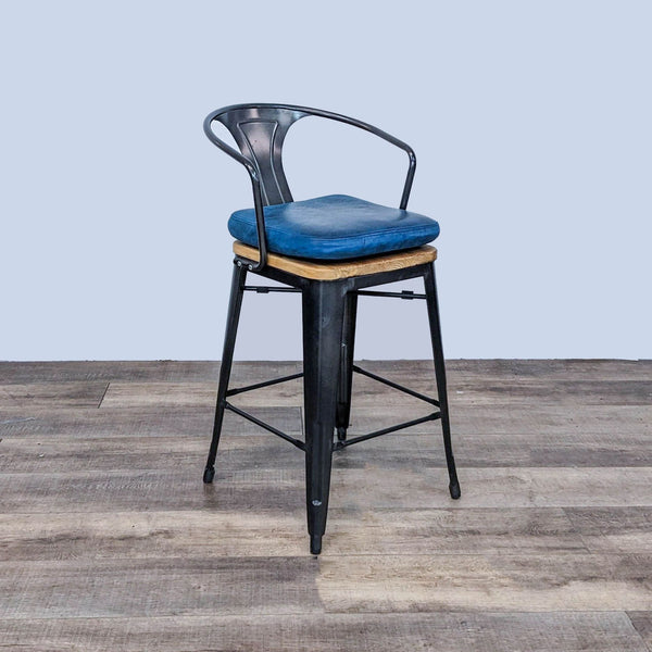 CB2 black metal frame barstool with a solid wood seat, removable blue cushion, on a wooden floor.