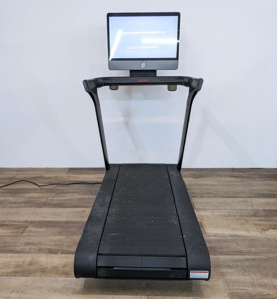 Front view of a Peloton treadmill with a touchscreen display on a wooden floor.