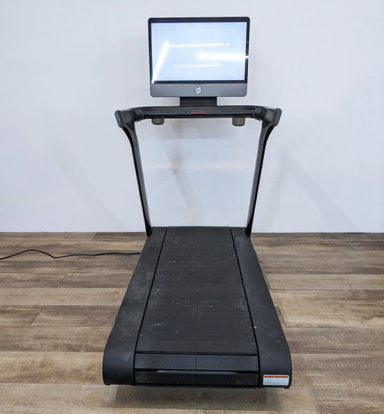 Front view of a Peloton treadmill with a touchscreen display on a wooden floor.