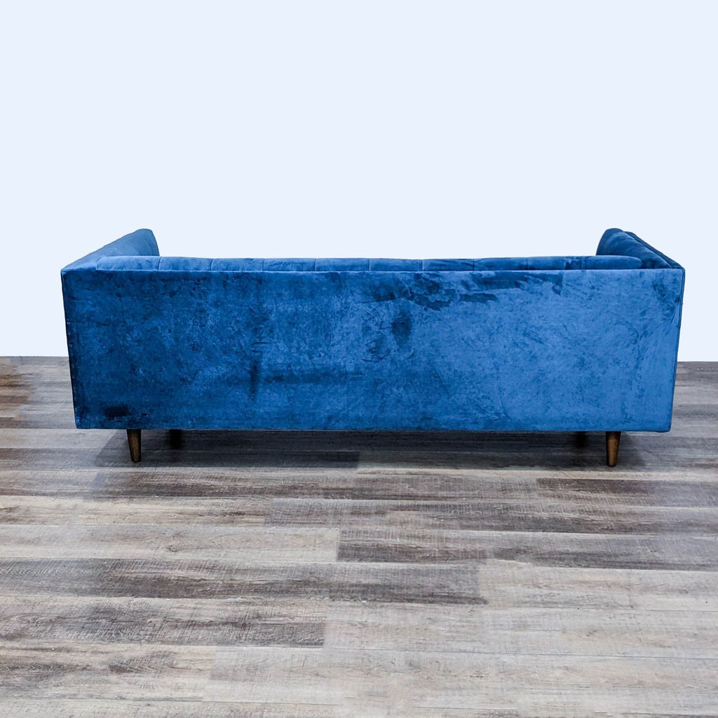 Rear view of a blue velvet sofa with tapered wood feet.