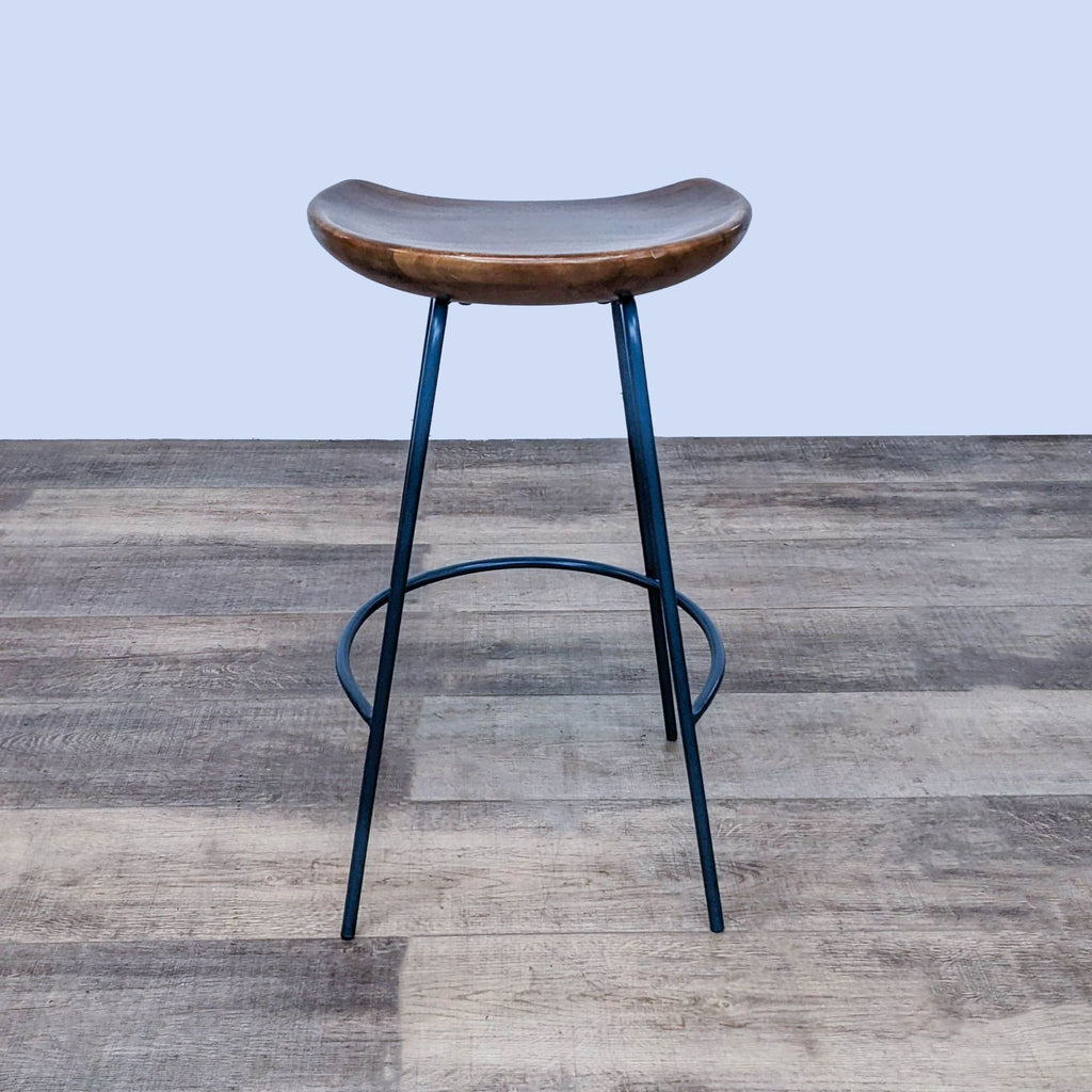 1. Counter stool with solid wood bucket seat and black steel base, designed by West Elm, shown on wood flooring.
