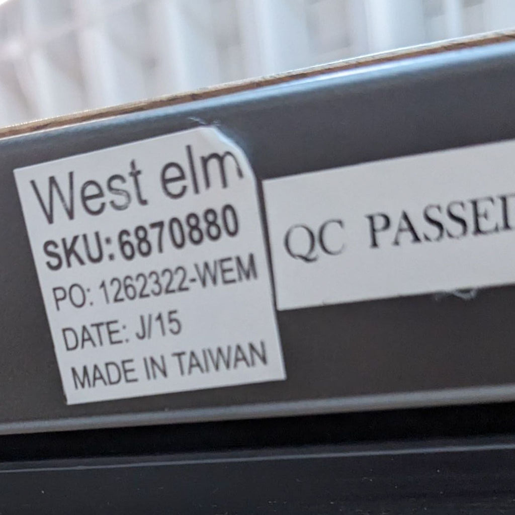 3. Close-up of a West Elm label on a metal base, indicating the Box Frame dining table's SKU, quality check passed, and manufacturing details.