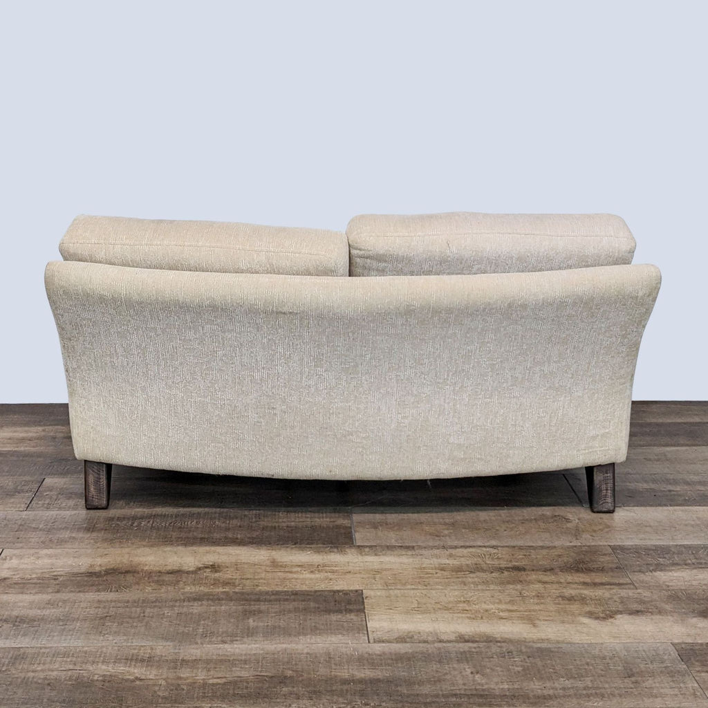 Rear view of a neutral beige Reperch loveseat with curvilinear back and wooden legs on a floor.
