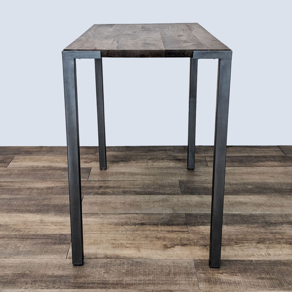 Stilt dining table by CB2, featuring a wood top and robust steel legs, photographed from a front angle.