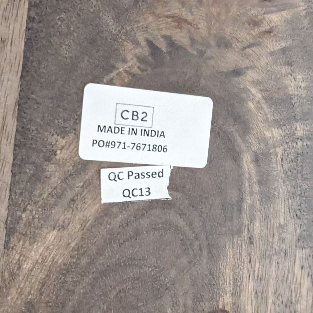 Close-up of a CB2 label on the Stilt dining table, indicating the brand, manufacturing origin, and quality control pass.