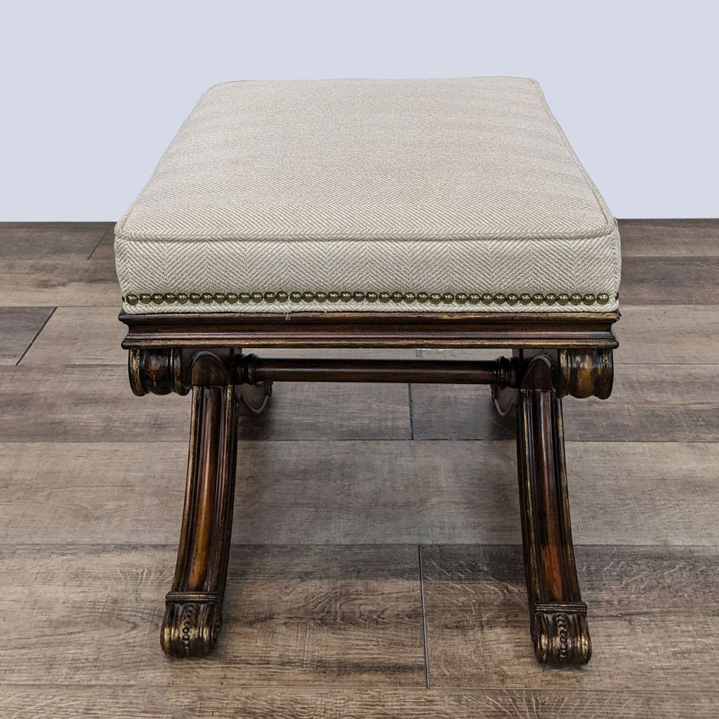 Carved wooden legged Ethan Allen ottoman with bronze accents and neutral upholstered top on a wood floor.