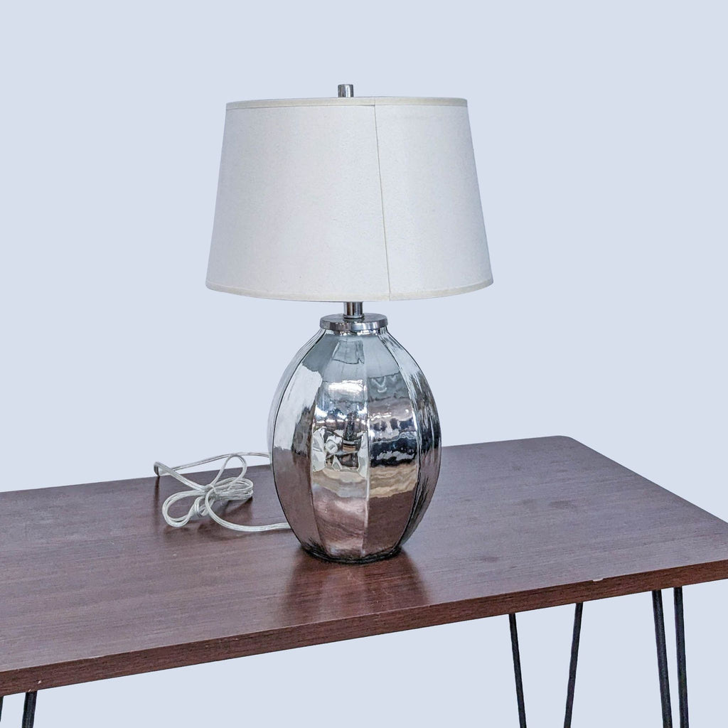 Pottery Barn silver table lamp with white shade on a wooden table, unplugged, on grey background.