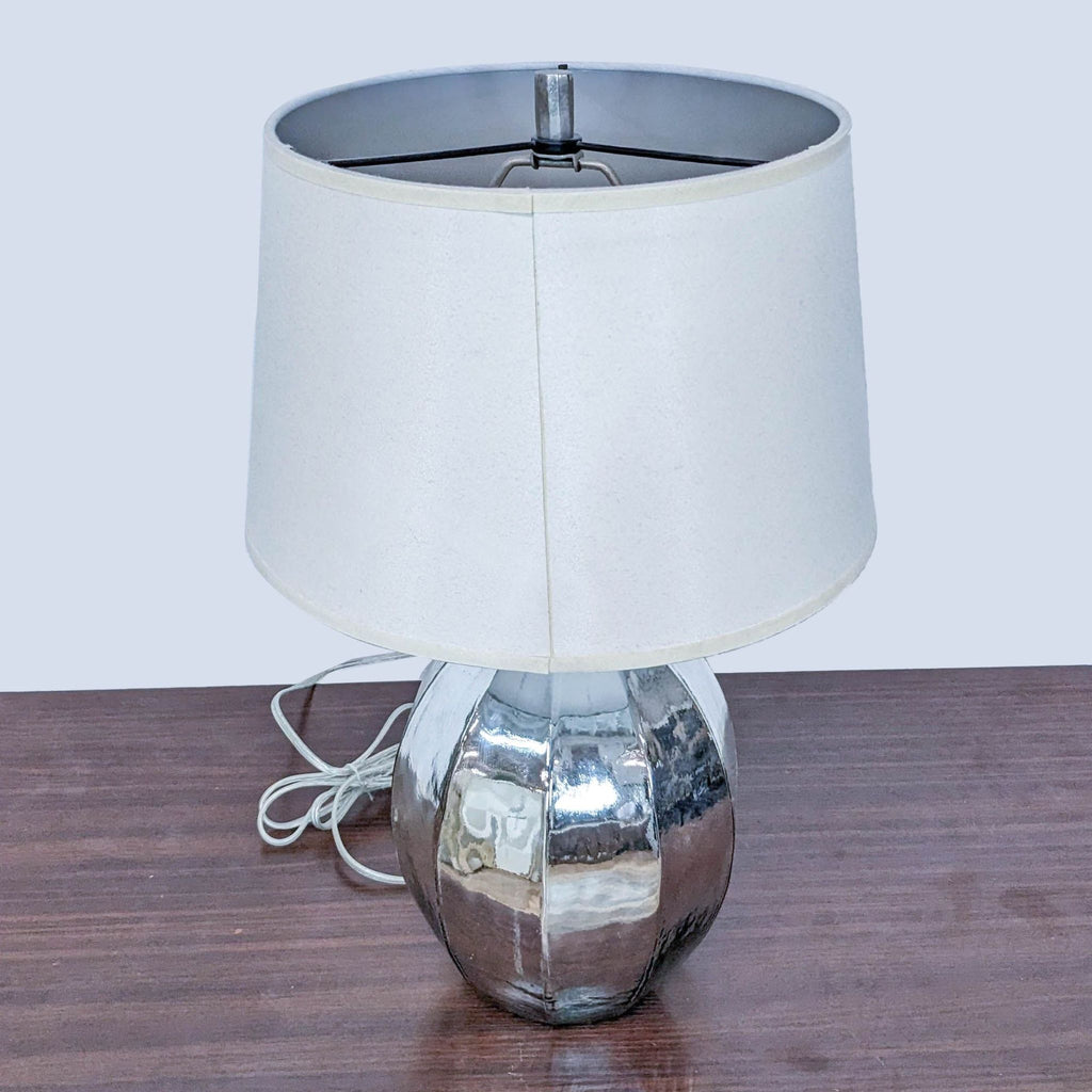 Close-up view of Pottery Barn reflective table lamp with off-white lampshade, on wooden surface.
