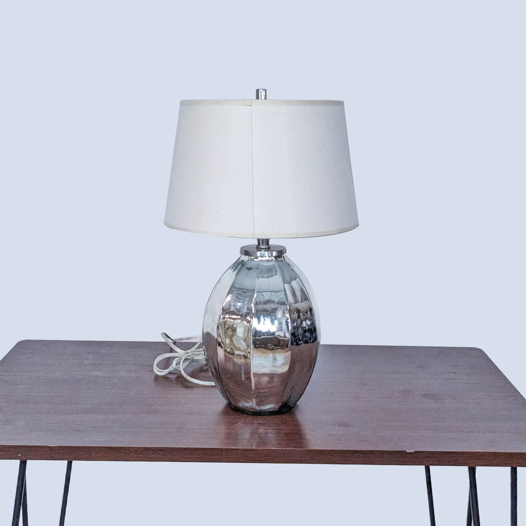 Silvery Pottery Barn lamp with a round base and cylindrical shade presented on a wooden table, cool tone backdrop.