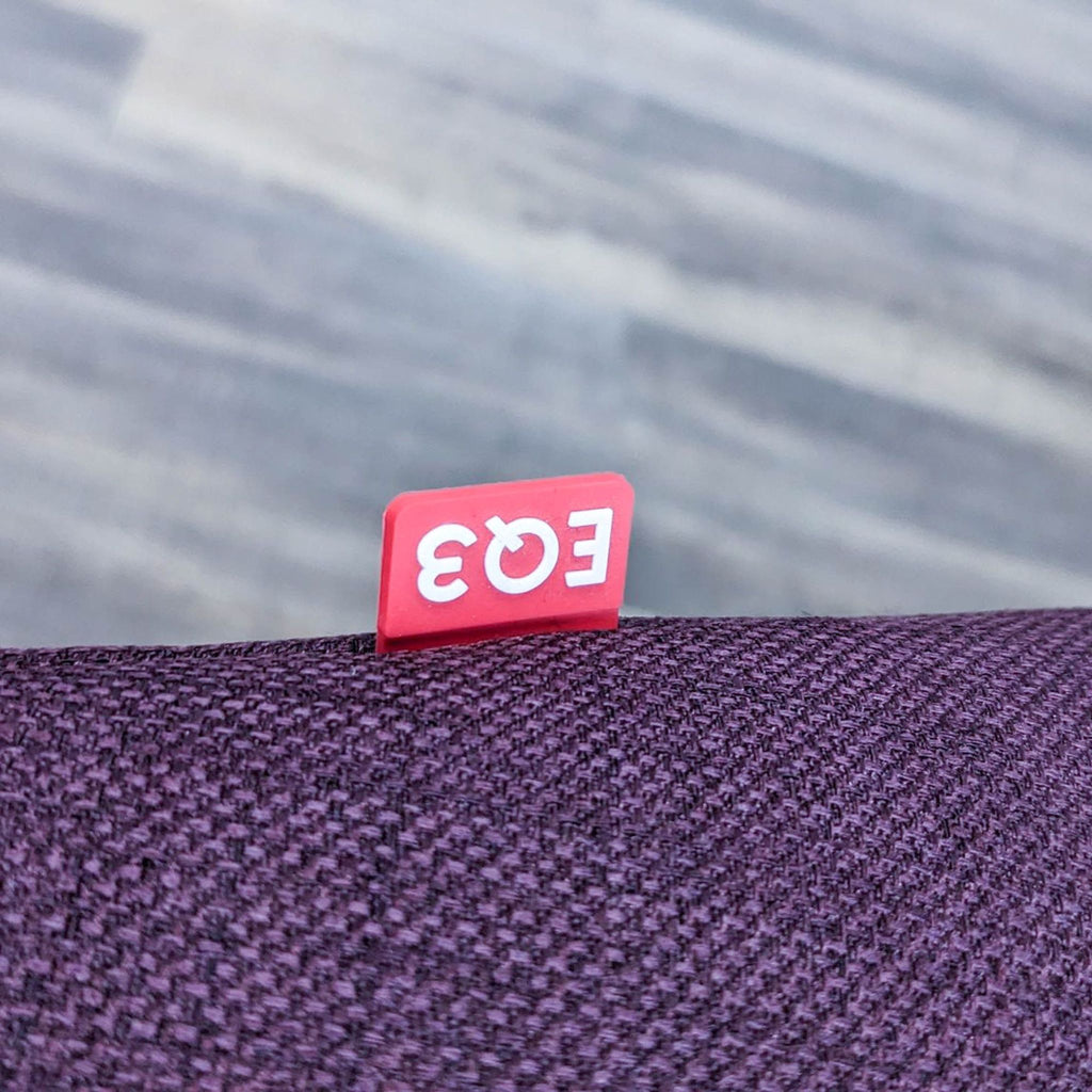 3. Close-up of the EQ3 logo tag on the Plum fabric upholstery of a modern lounge chair with textured material.