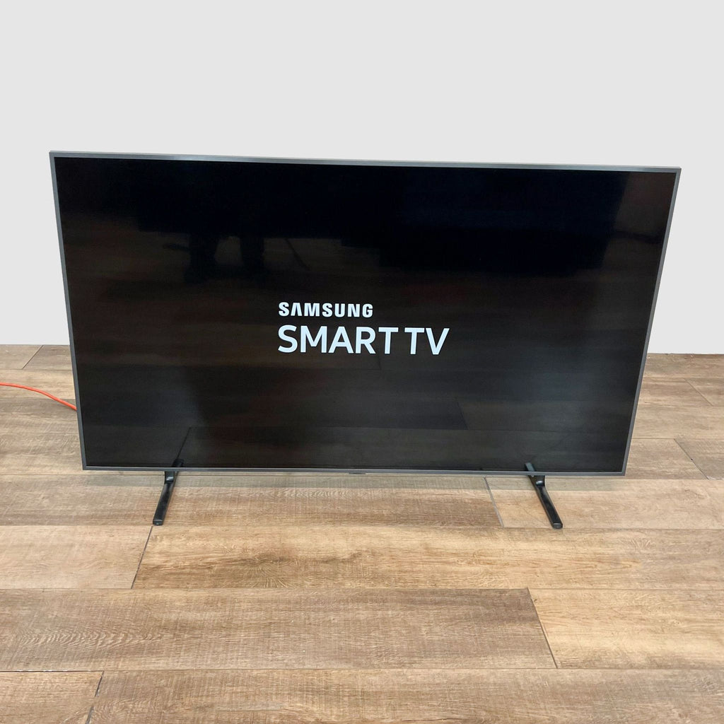 Front view of a Samsung Smart TV featuring a sleek black screen with the brand logo, perched on a minimal stand against a wooden floor.