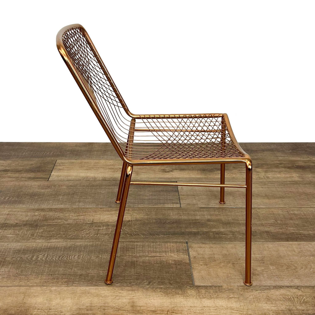 Brass-finished CB2 Modern Beta dining chair with graphic linear wire design over a wooden floor, showcasing profile view.