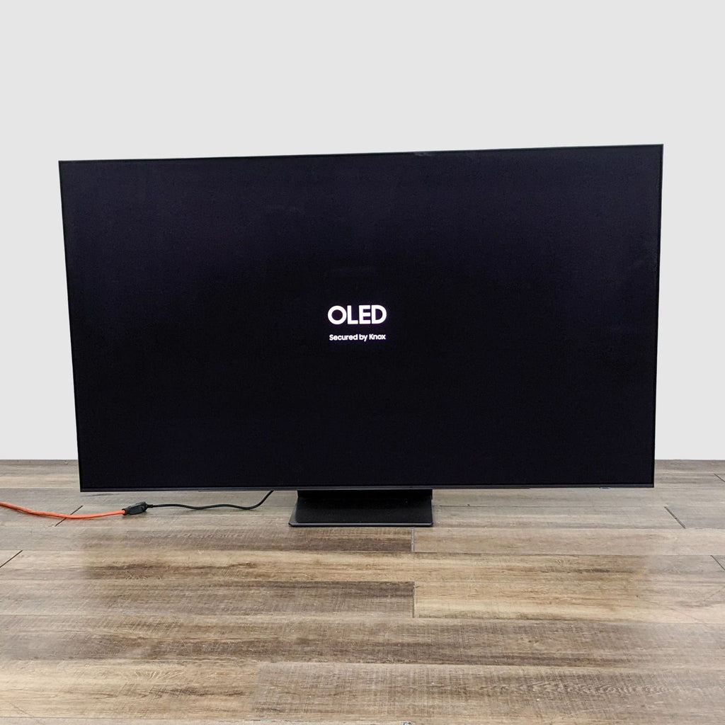 A modern Samsung OLED TV secured by Knox, featuring brilliant picture quality, on a wooden floor.
