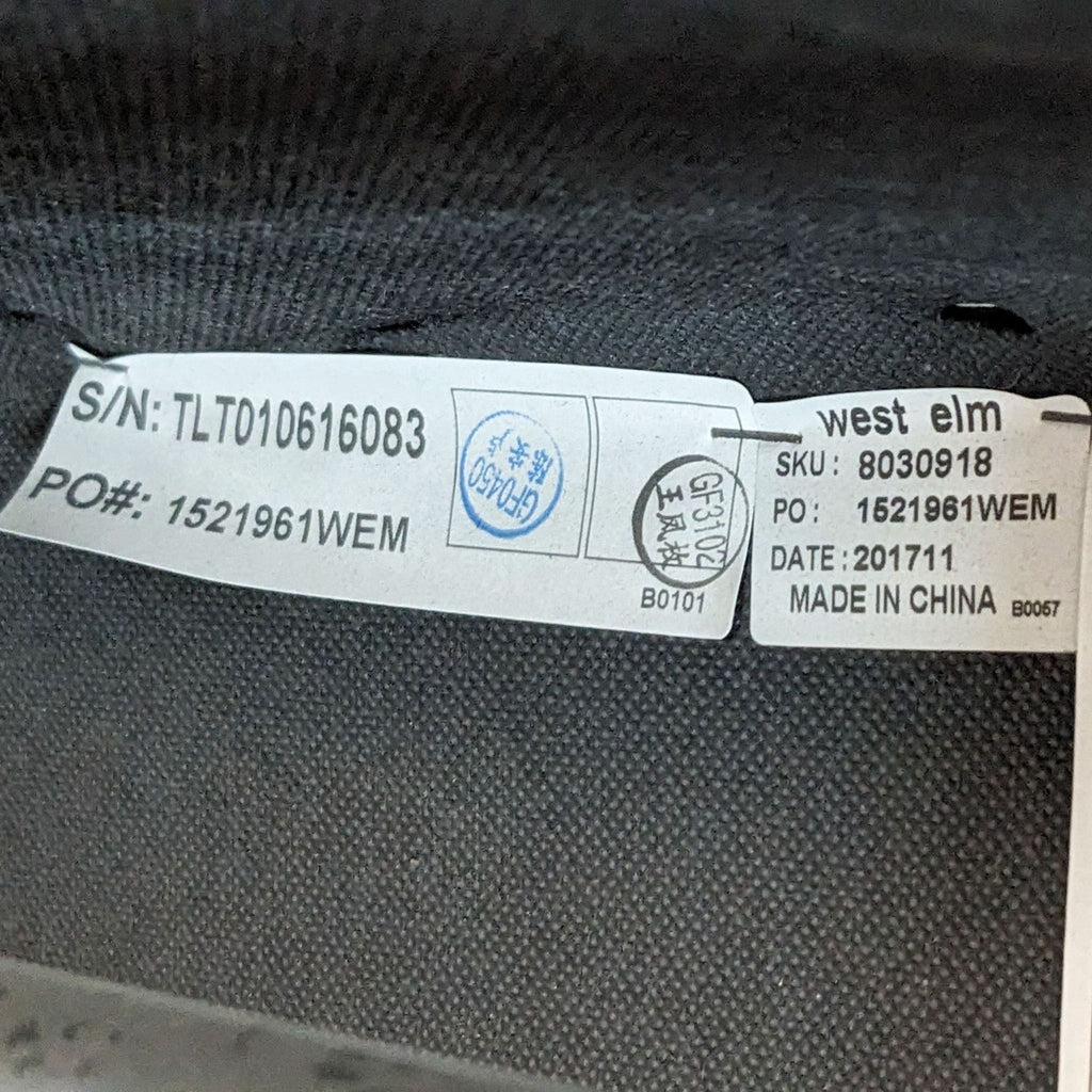 3. "Close-up of a West Elm label on a gray fabric ottoman, indicating brand, SKU, and manufacturing details."