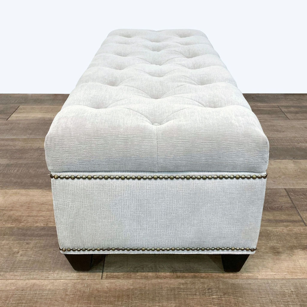 Beige tufted ottoman bench with wooden feet on a hardwood floor, viewed from the side.