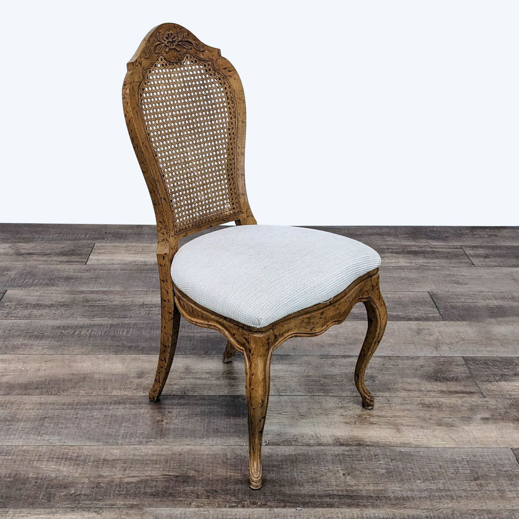 3. Angled view of an upholstered Reperch Louis XV dining chair with intricate wood carving and cane back against wood flooring.