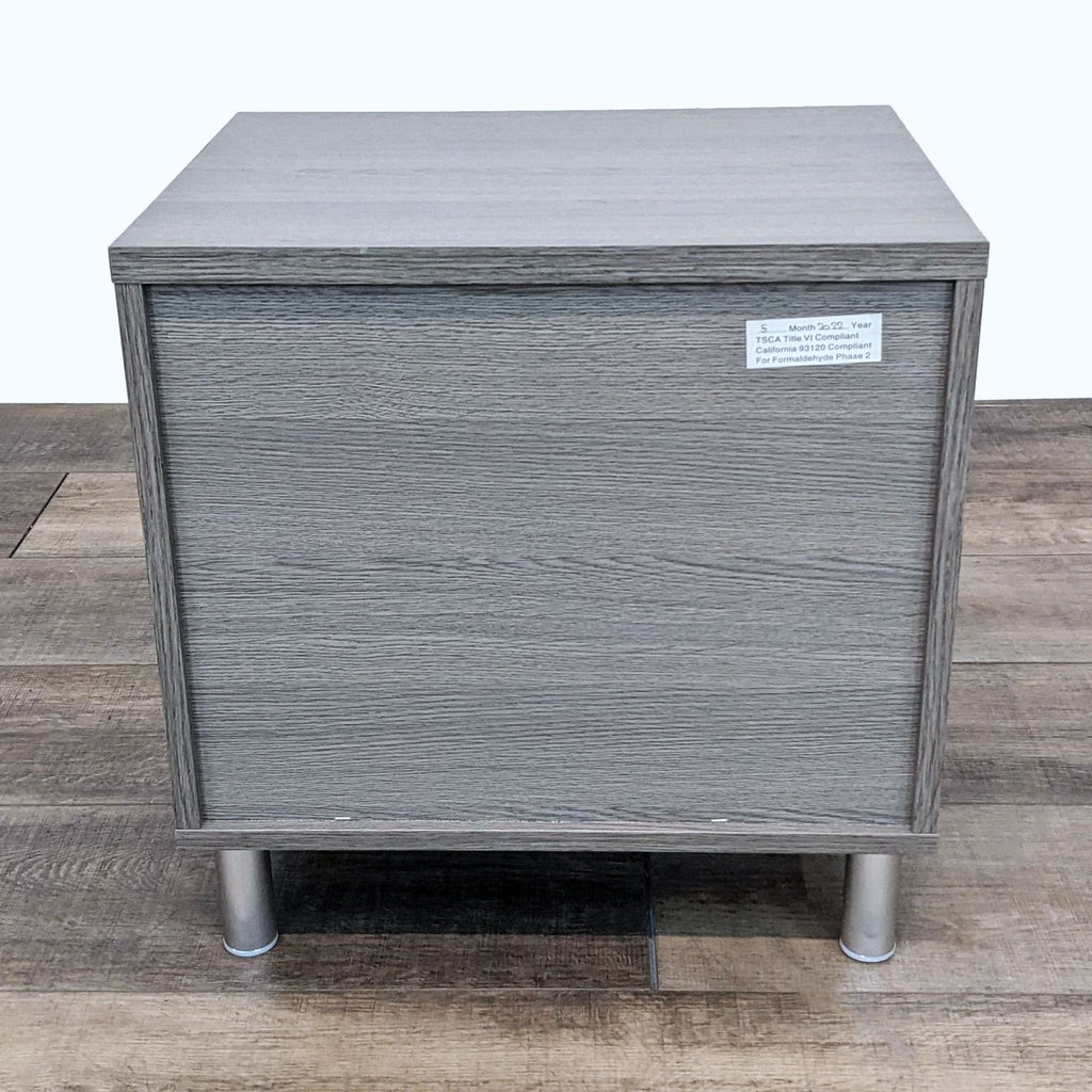 3. Grey wooden end table with a closed top drawer showing a compliance label, by Ana Furniture.