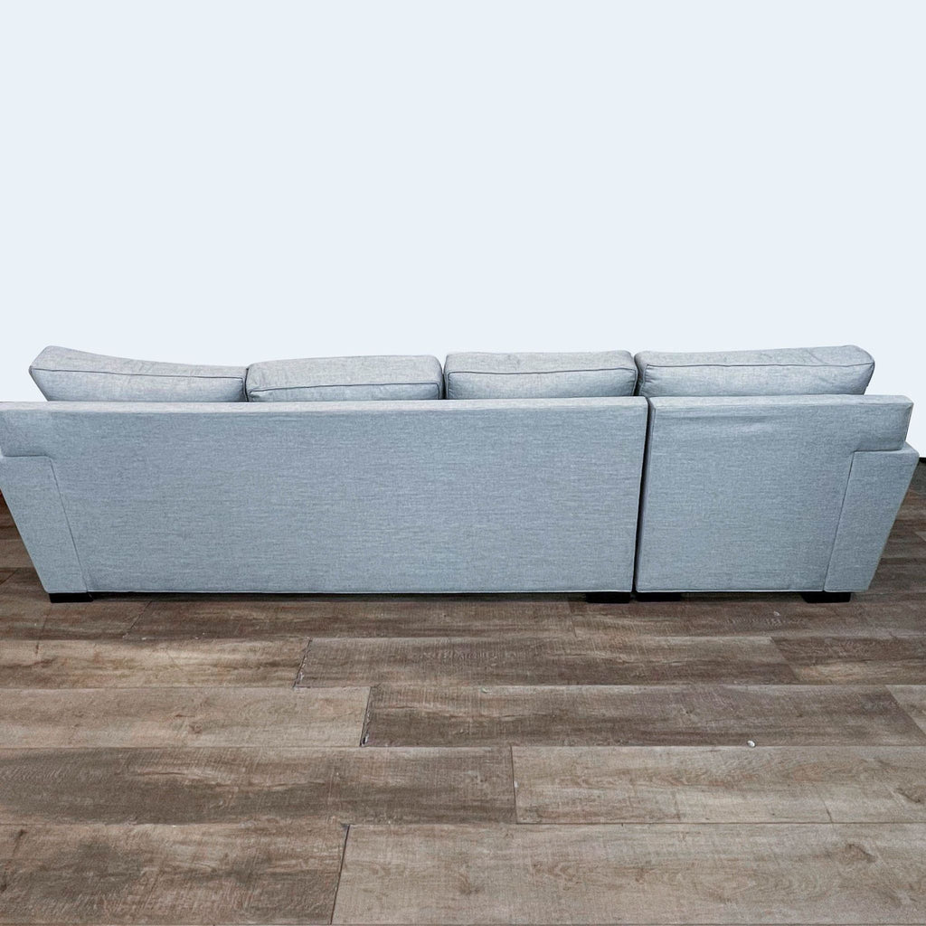 Rear view of a light gray upholstered Stanton sectional sofa with detachable chaise on a wooden floor against a white wall.
