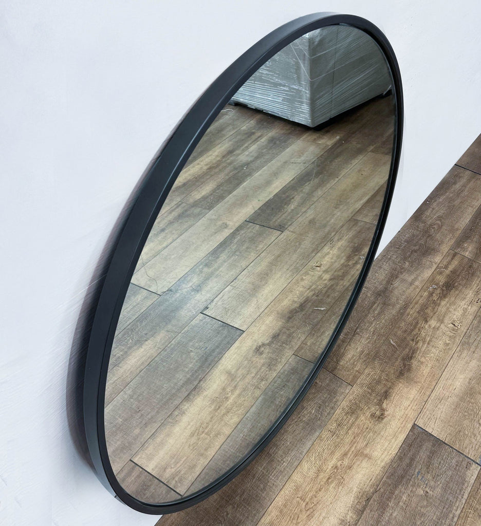 Leaning round mirror by Williams-Sonoma with a metal edge, reflecting wooden flooring.