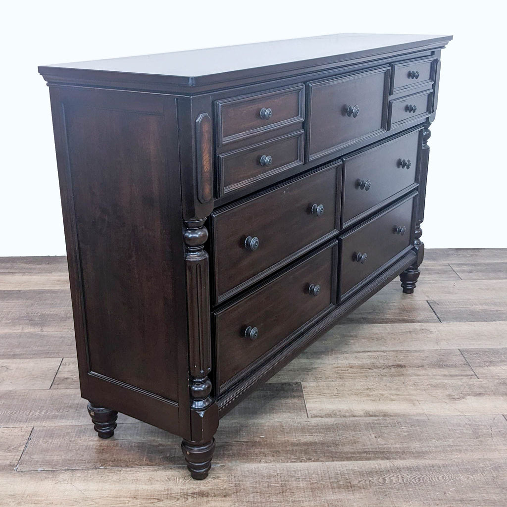 Front-angle perspective of an ornate Ashley Signature dresser with seven drawers and dark bronze colored hardware on a wood floor.