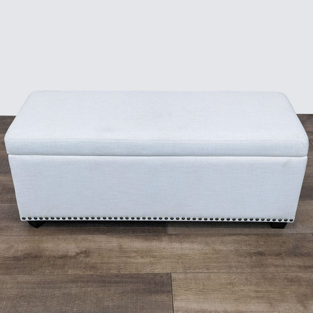 Neutral linen upholstered storage ottoman by Living Spaces, featuring nailhead detailing and dark wood legs.