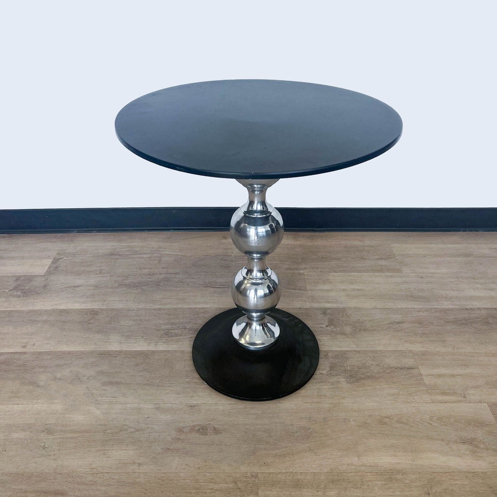 A polished metallic spindle supports a circular black tabletop, Reperch end table against a white wall.