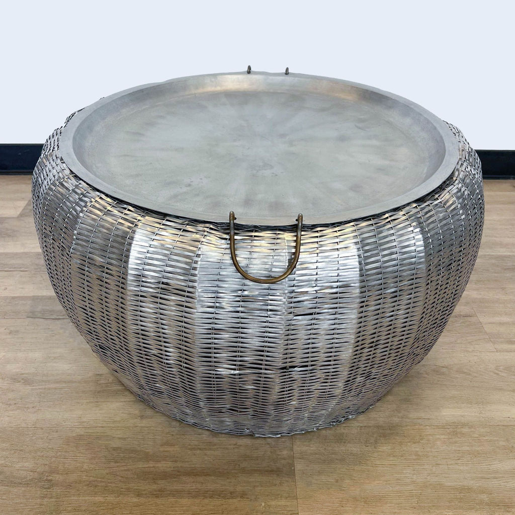 3. Round metal coffee table in a basket weave design with a removable top, part of the Reperch collection, on a wooden floor with a grey wall behind.