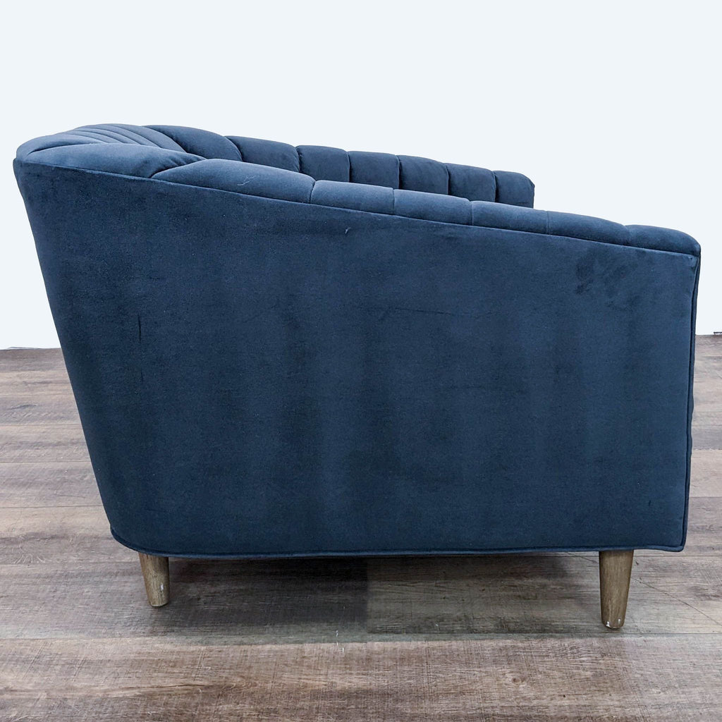 Rear view of a Four Hands lounge chair in blue velvet with tufted barrel back and angled wooden legs.