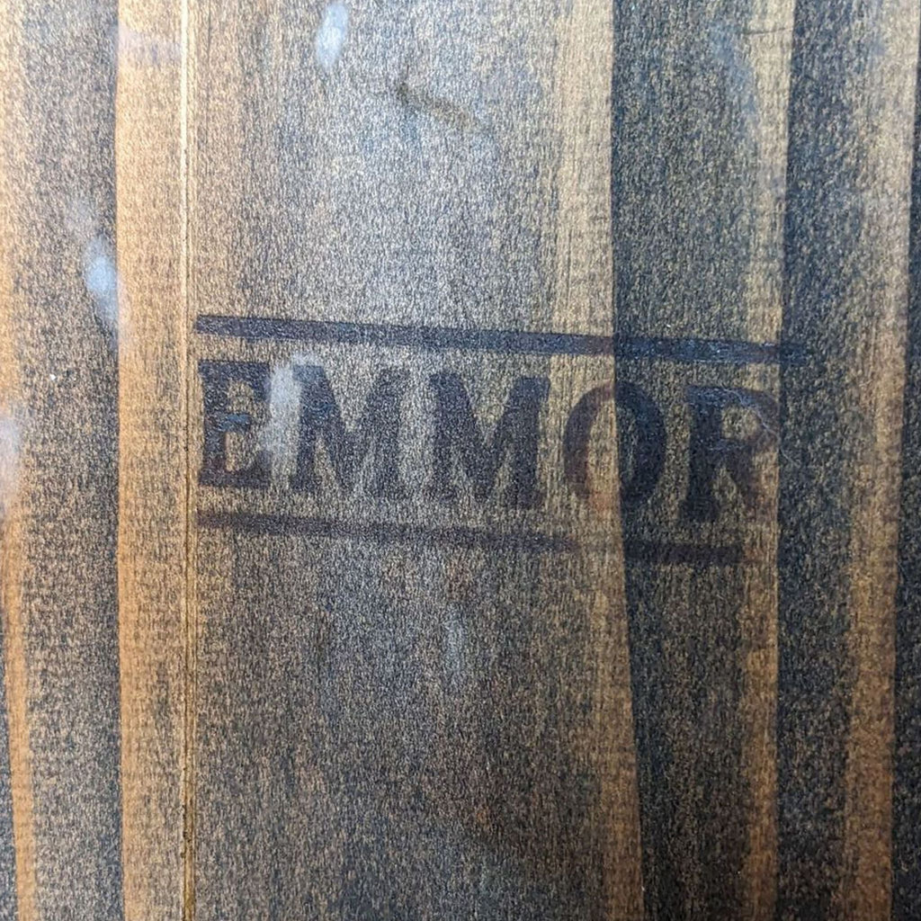 3. Close-up of the natural grained wood of an Emmor dining bench, featuring the brand's name stamped on the surface.