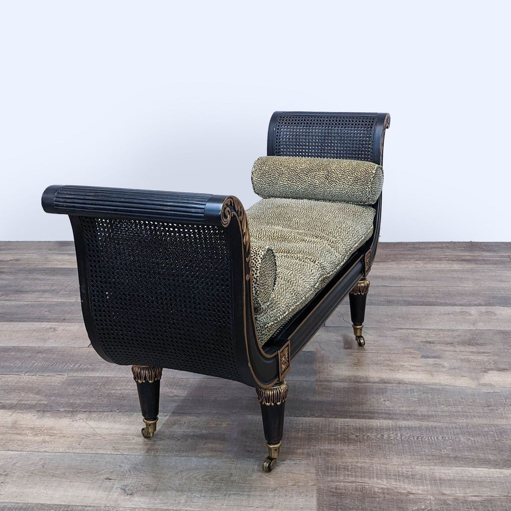 Angle view of Empire 59" black bench by Reperch with cane seating, ornate gold detail, and two bolsters on wood floor.