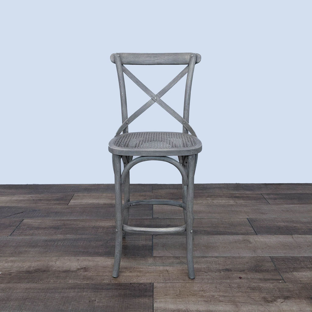 Single grey barstool with an X-shaped back and cane seat on a wooden floor.
