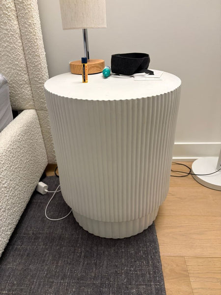 1. White ribbed cylindrical West Elm end table beside a bed with a lamp, eye mask, and small items on top.