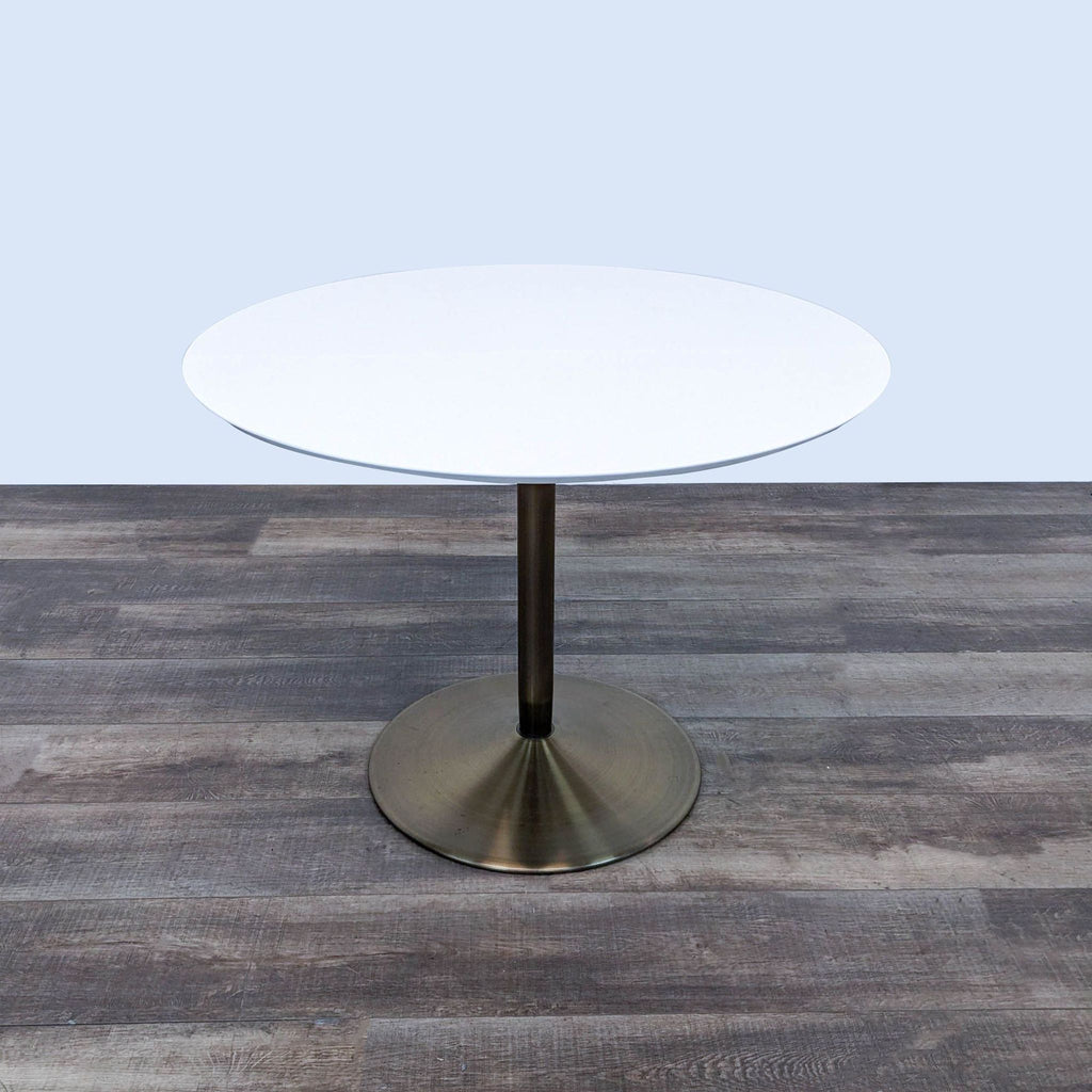 Round dining table from CB2 with sleek white top and brass metal pedestal, placed on wood floor.  