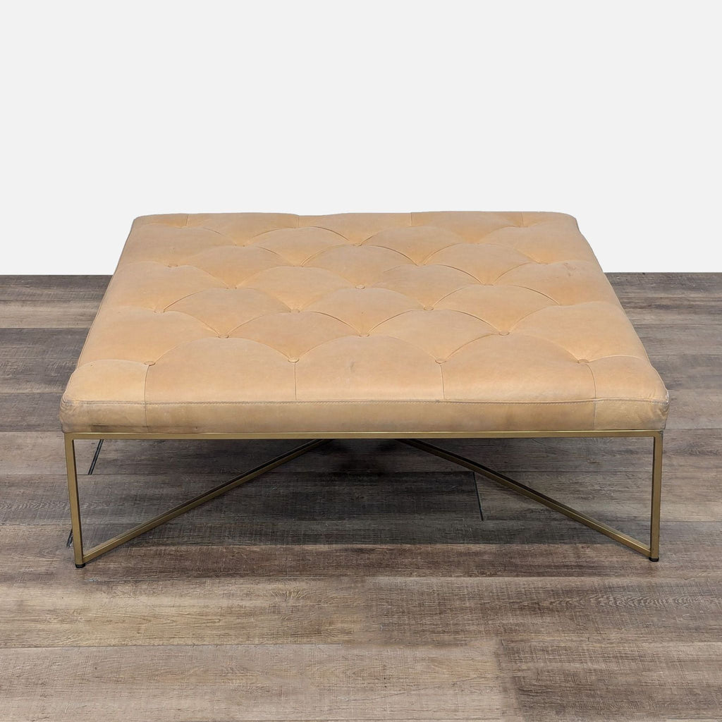 a beige leather ottoman with a gold metal base.