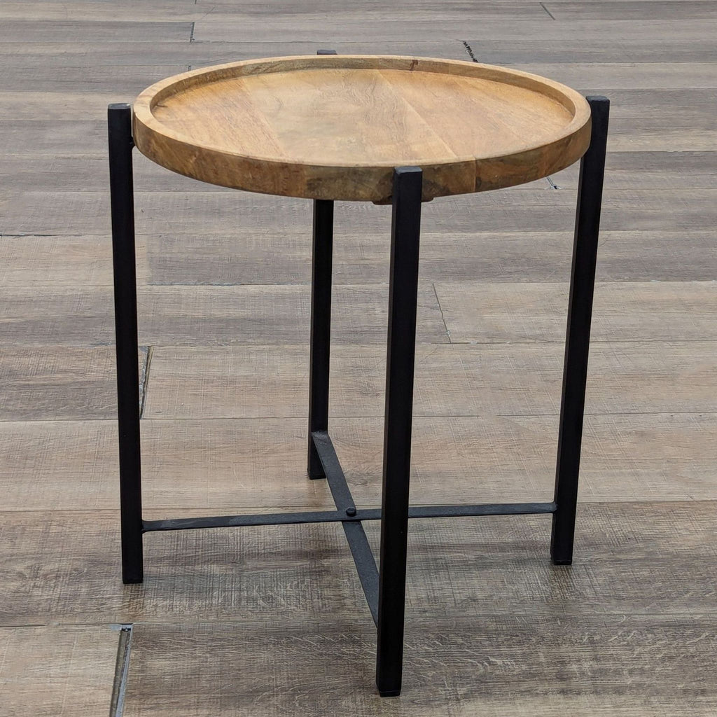 Round wooden side table with black metal legs on a wooden floor.