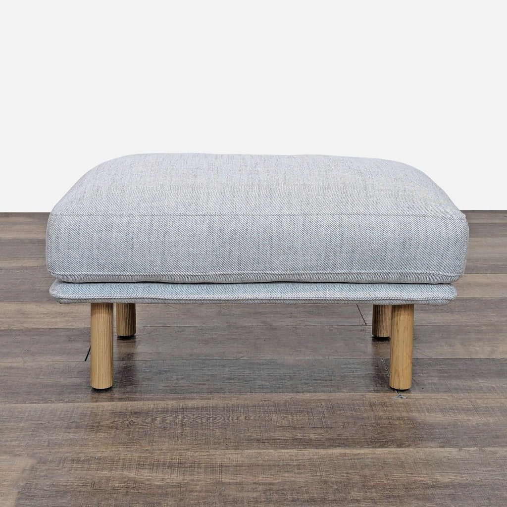 Gray ottoman with wooden legs on a wood floor, seen from the front.