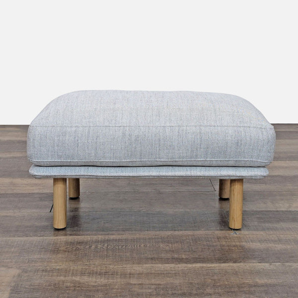 Gray ottoman with wooden legs on a wood floor, seen from the front.
