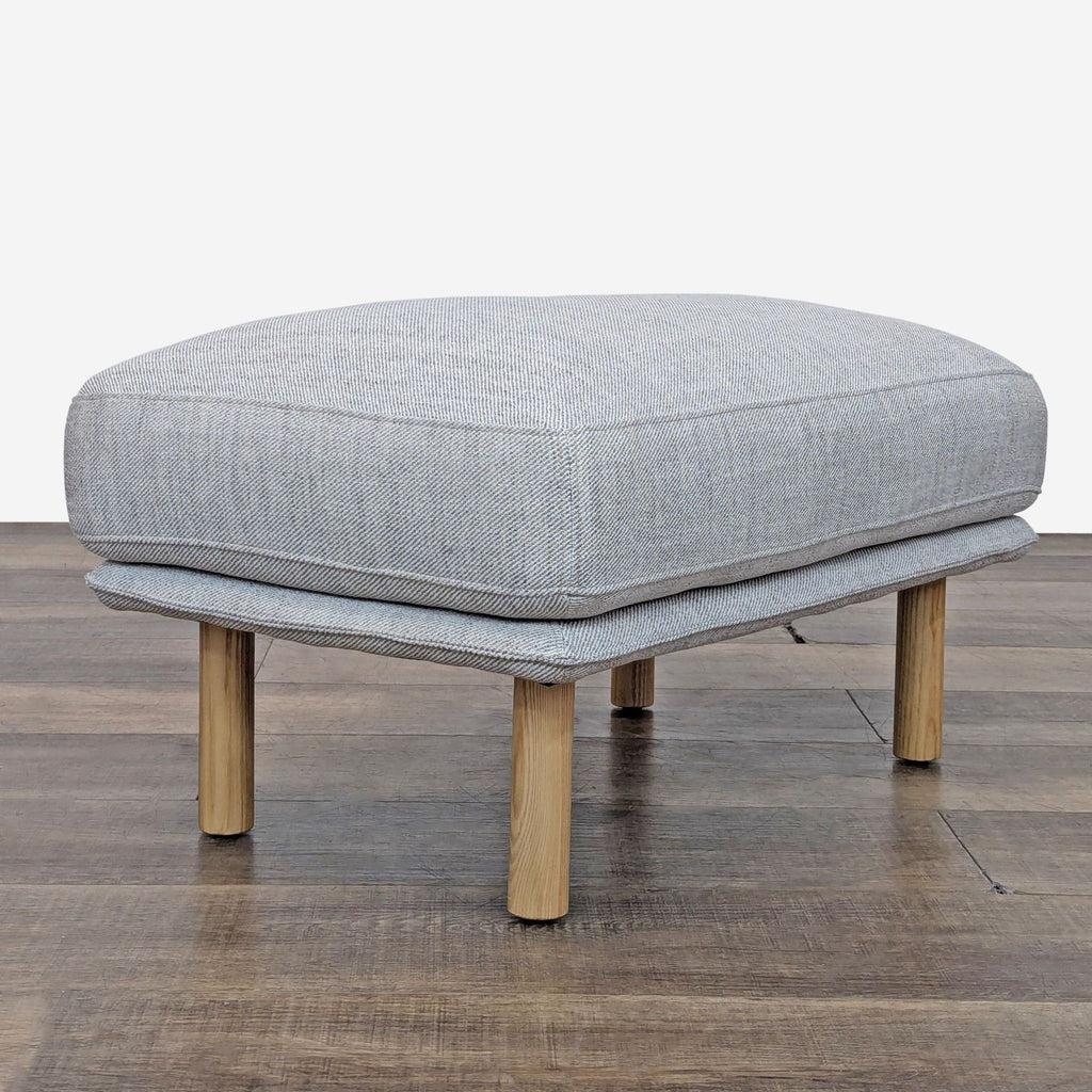Side view of a gray ottoman with wooden legs on a wooden floor.