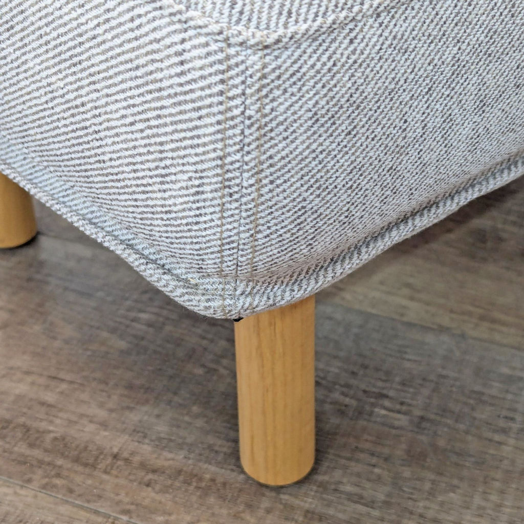 Close-up of the corner of a gray ottoman, showing texture and wooden leg.