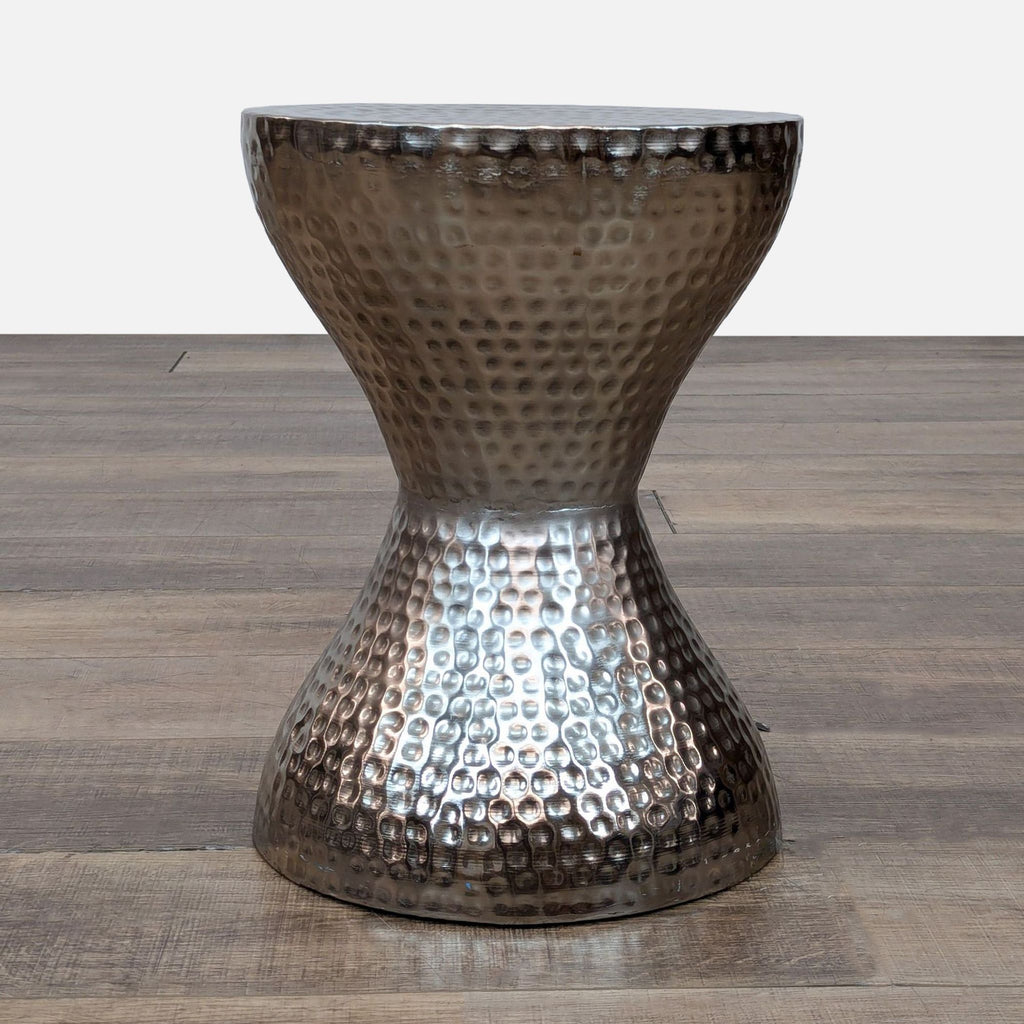 Hammered metal side table with an hourglass shape on a wooden floor.