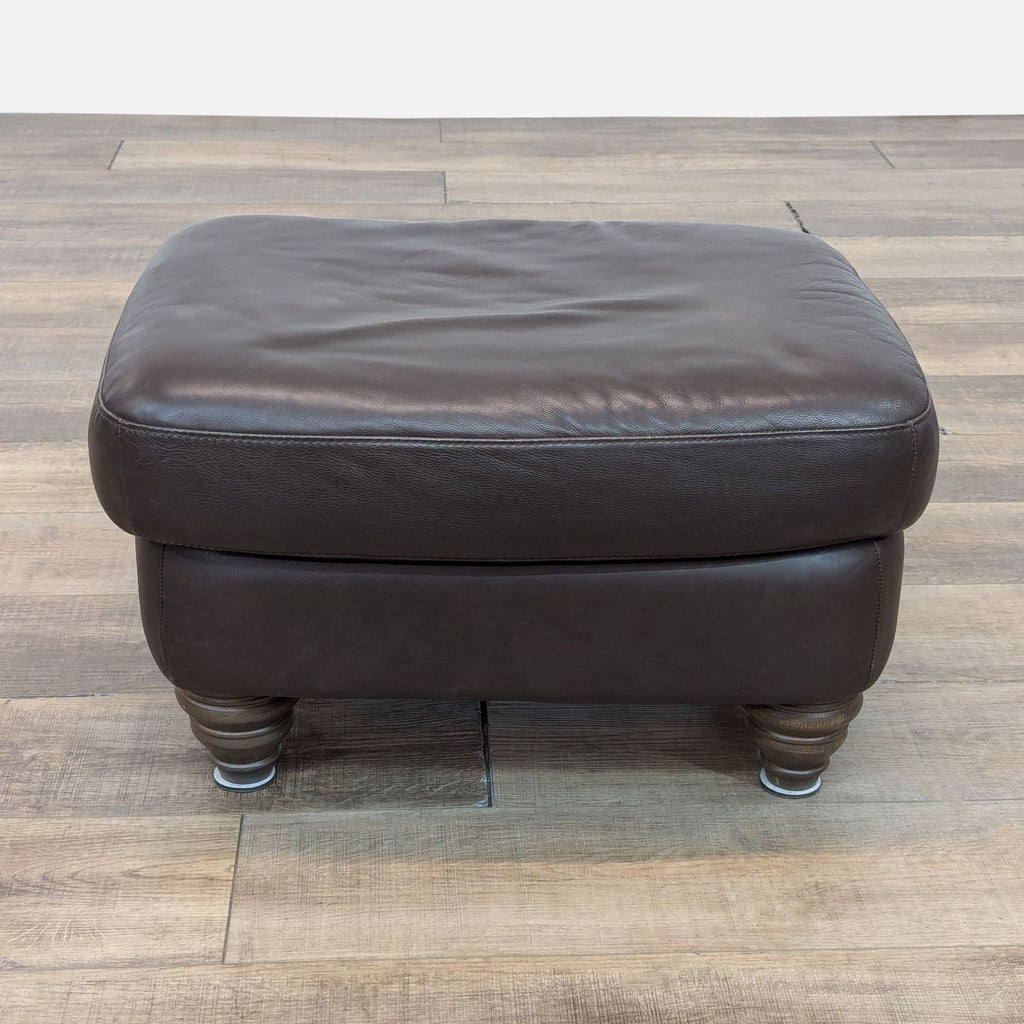 Brown leather ottoman with turned wood bun feet, seen at an angle on wooden flooring.